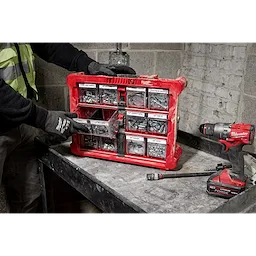 A person wearing gloves organizes a red portable tool organizer box containing various screws and fasteners. The box is placed on a workbench next to a Milwaukee cordless drill and a hammer. The background features a concrete and brick wall.