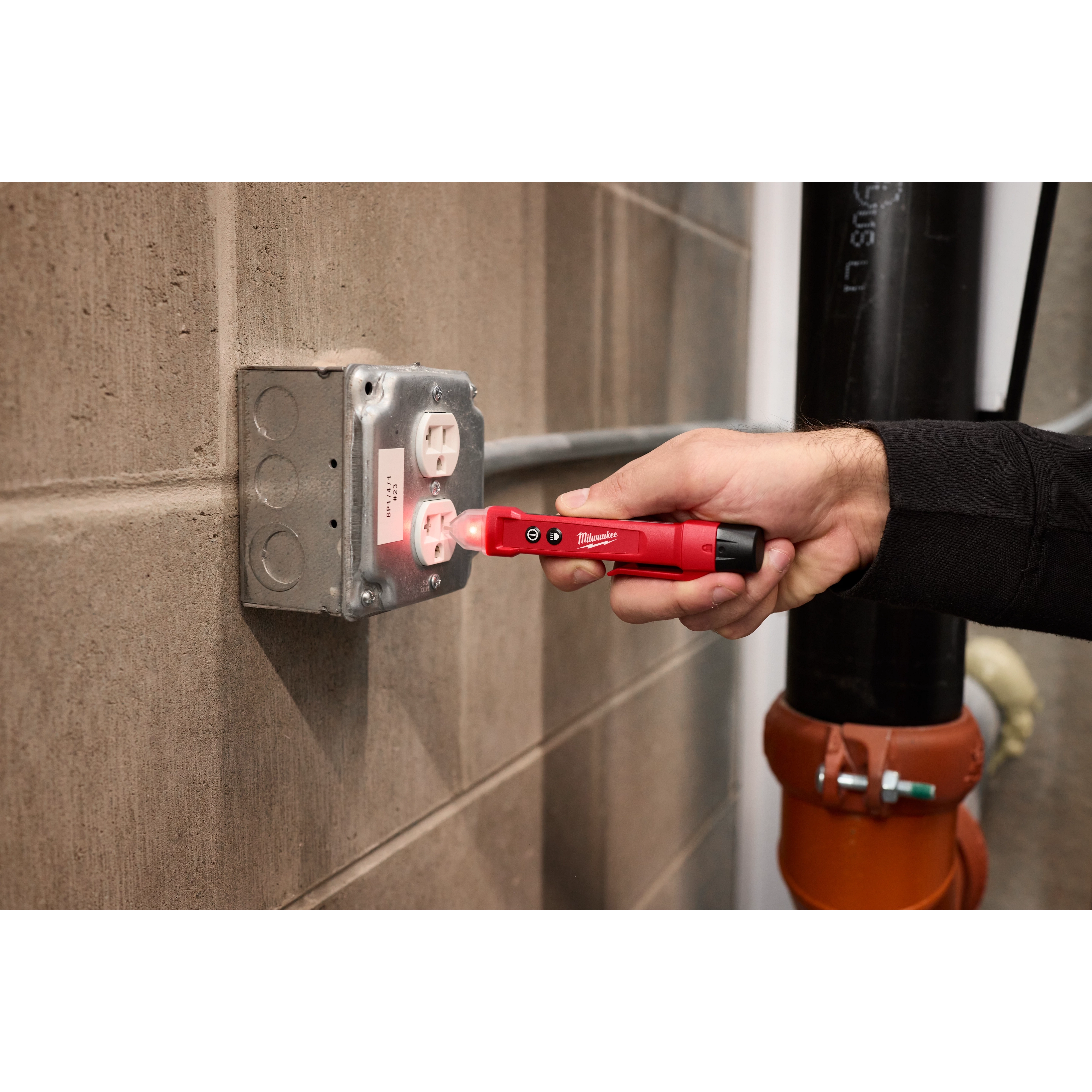 A person is using the Voltage Detector w/ LED to test a wall outlet. The handheld tool emits a red light and the user is holding it against the outlet, which is mounted on a concrete wall.