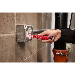 A person is using the Voltage Detector w/ LED to test a wall outlet. The handheld tool emits a red light and the user is holding it against the outlet, which is mounted on a concrete wall.
