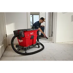 The M18 FUEL™ NEXUS™ Dual Battery Wet/Dry Vacuum Motor Head with PACKOUT™ Compatibility & VACLINK™ is shown in use on a job site. The vacuum is red with black accents and connected to a flexible hose, efficiently cleaning debris from the floor.