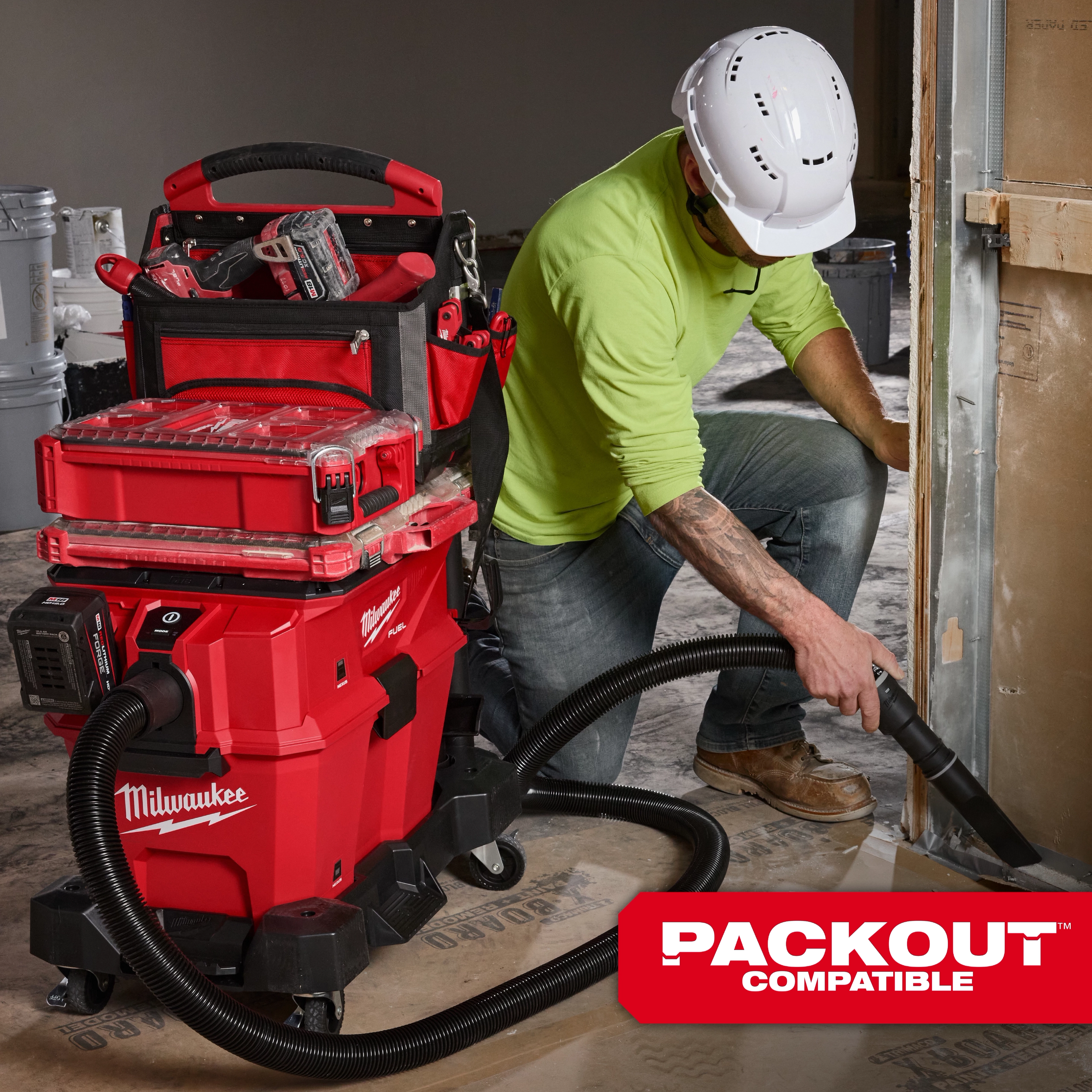 A worker uses the M18™ FUEL™ NEXUS™ 6 Gallon Wet/Dry Vacuum with PACKOUT™ Compatibility, featuring a red body, multiple storage compartments, and a flexible hose attachment. The vacuum is positioned near construction materials, and the worker is wearing a white hard hat and green shirt.