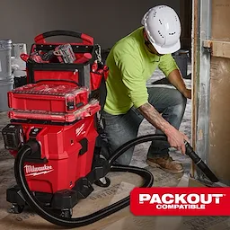 A worker uses the M18™ FUEL™ NEXUS™ 6 Gallon Wet/Dry Vacuum with PACKOUT™ Compatibility, featuring a red body, multiple storage compartments, and a flexible hose attachment. The vacuum is positioned near construction materials, and the worker is wearing a white hard hat and green shirt.