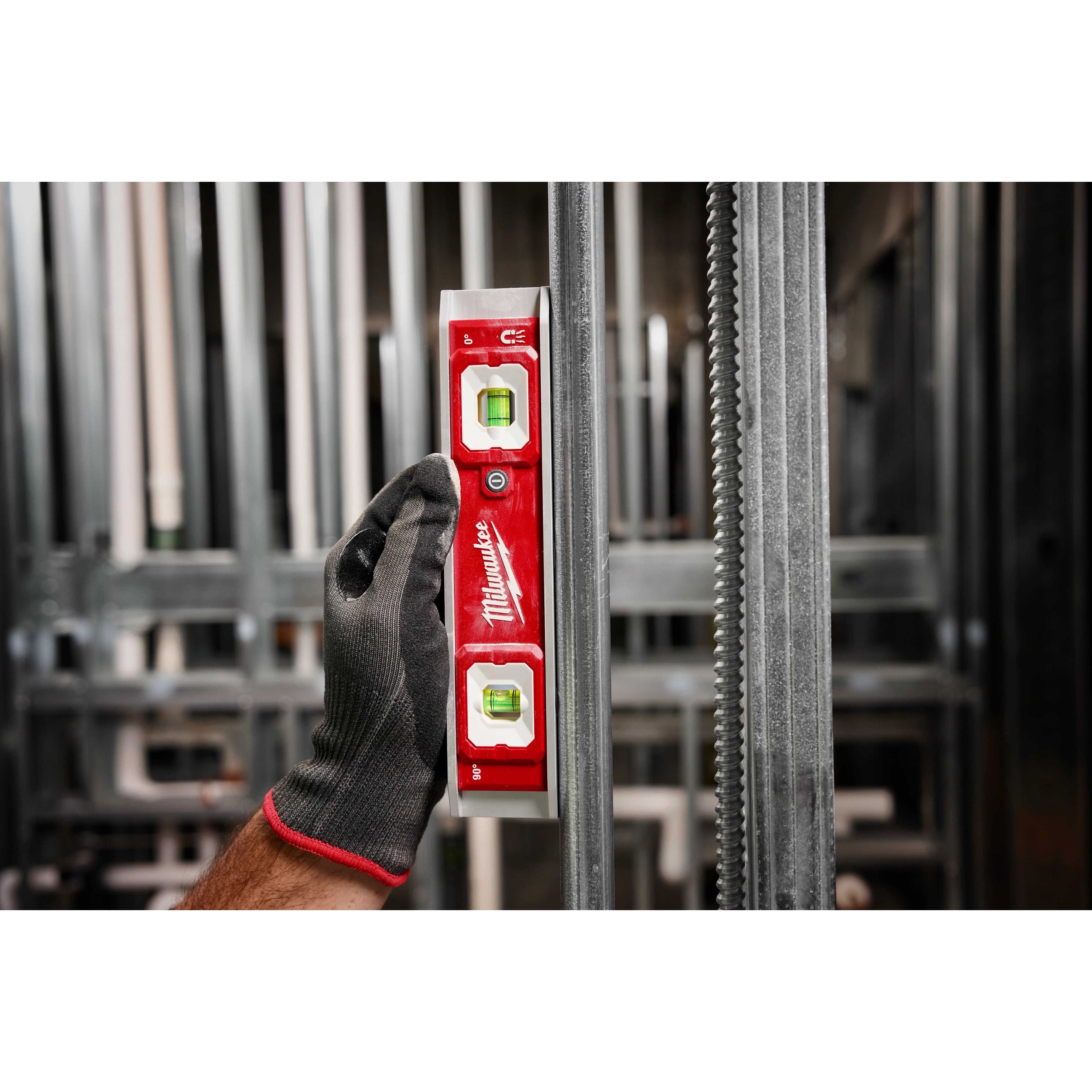 A gloved hand holds a 9" Torpedo Level with Lighted Vials against a vertical metal surface. The tool is red with two illuminated vials for accurate leveling. The background features a blurred construction setting with metal beams and frames.
