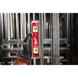 A gloved hand holds a 9" Torpedo Level with Lighted Vials against a vertical metal surface. The tool is red with two illuminated vials for accurate leveling. The background features a blurred construction setting with metal beams and frames.