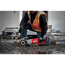 A person in a safety vest and gloves is using a Milwaukee M18 FUEL™ Pipe Threader with ONEKEY on a gravel surface, next to industrial equipment.