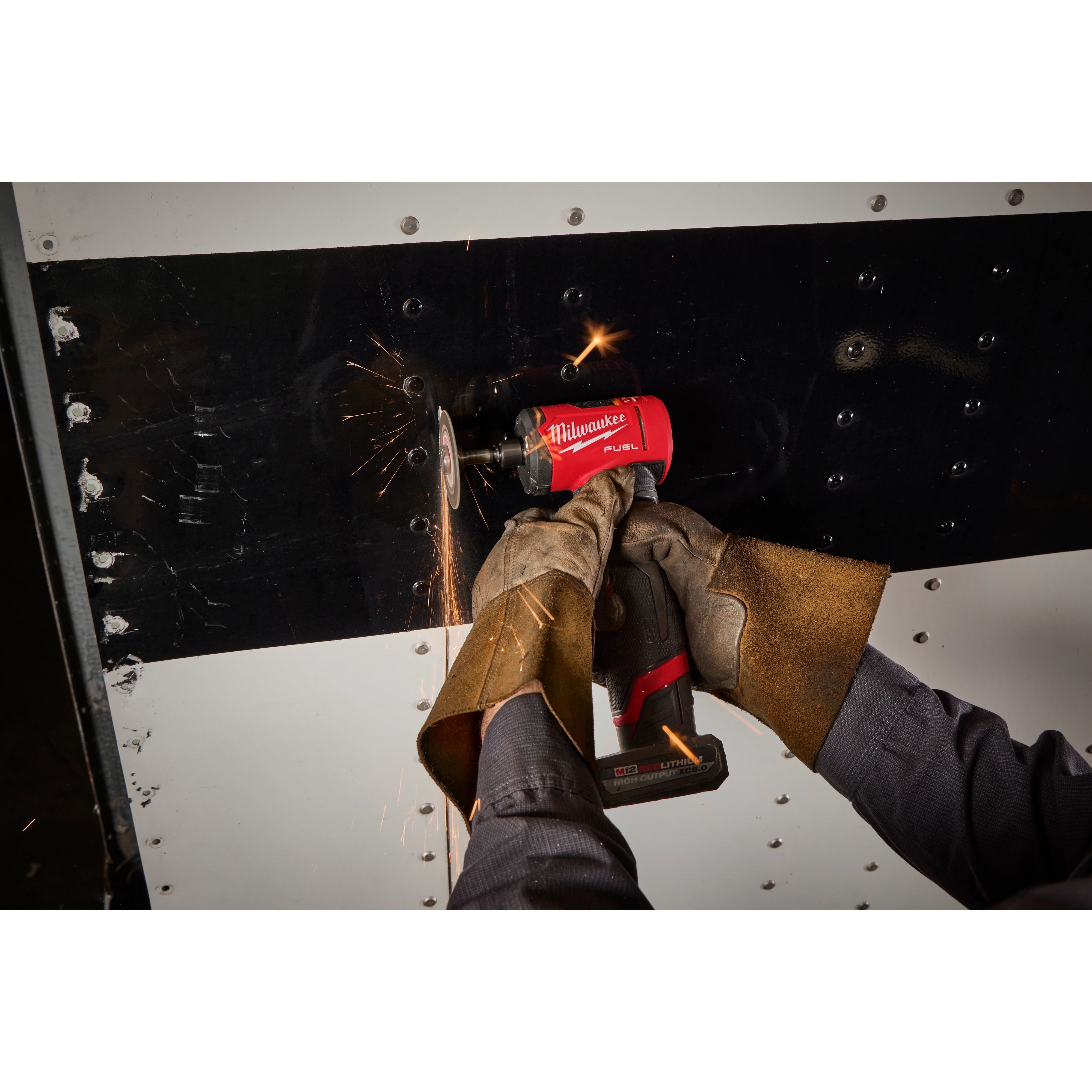 A person uses an M12 FUEL™ 1/4" Right Angle Die Grinder on a metal surface, producing sparks. The tool is being operated with both hands, wearing brown protective gloves. The scene highlights the grinder’s use for cutting or grinding tasks.