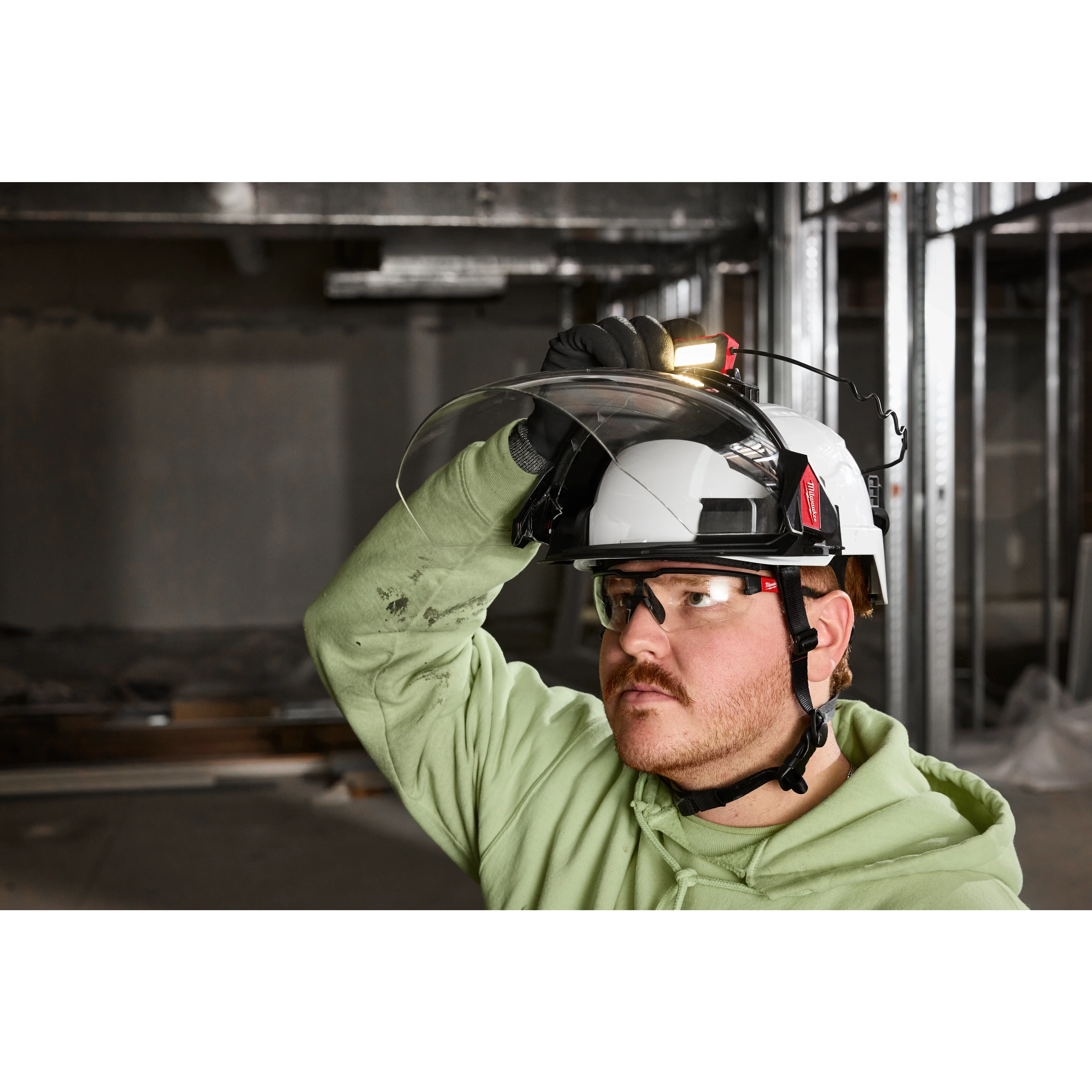 A person wearing a BOLT™ Gasketed Full Face Shield - Clear Dual Coat Lens (Compatible with Milwaukee® BOLT™ Safety Helmets & BOLT™ Hard Hats) on a white safety helmet, inside a construction site.