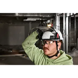 A person wearing a BOLT™ Gasketed Full Face Shield - Clear Dual Coat Lens (Compatible with Milwaukee® BOLT™ Safety Helmets & BOLT™ Hard Hats) on a white safety helmet, inside a construction site.