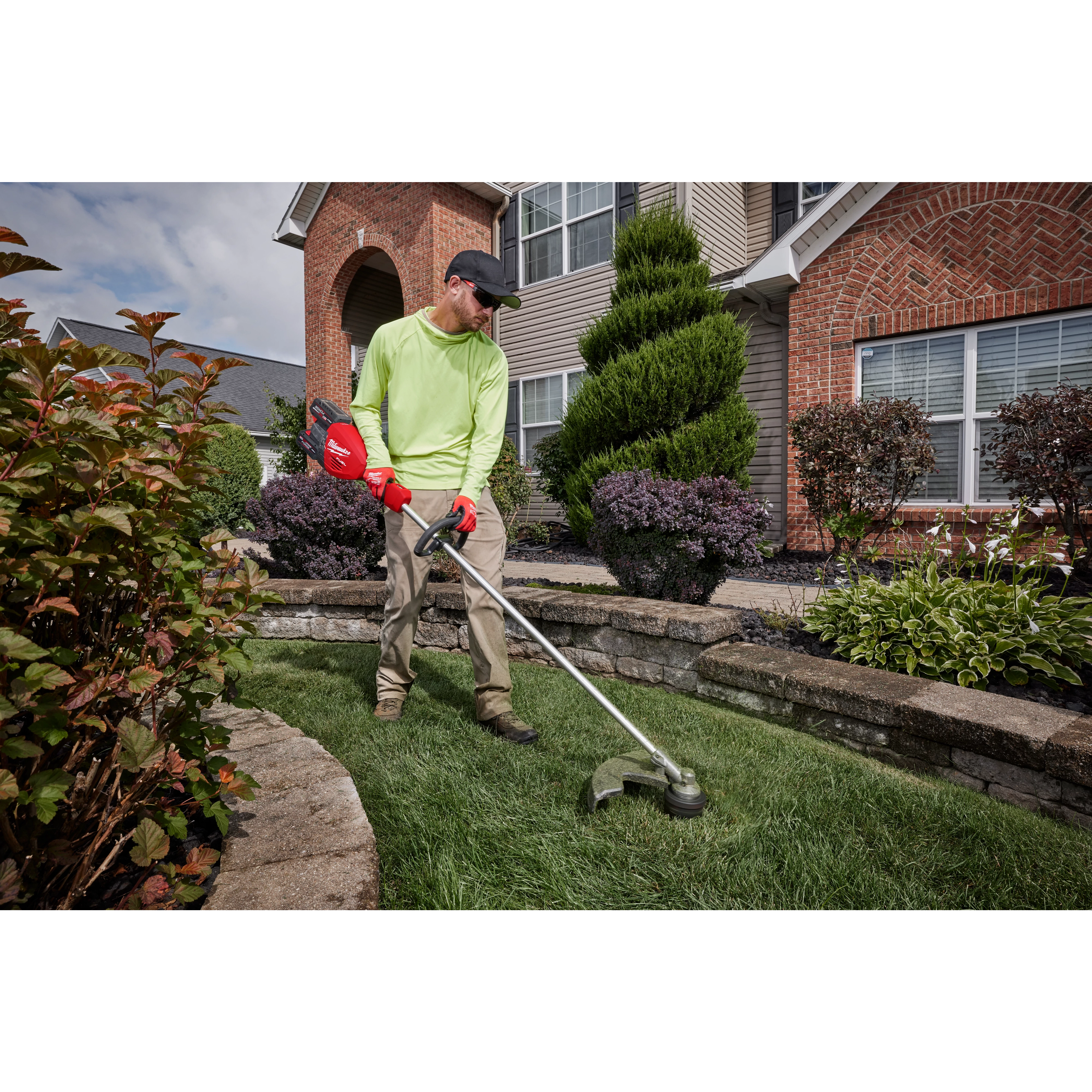 Person using a string trimmer to edge the lawn in front of a house surrounded by neatly landscaped plants.