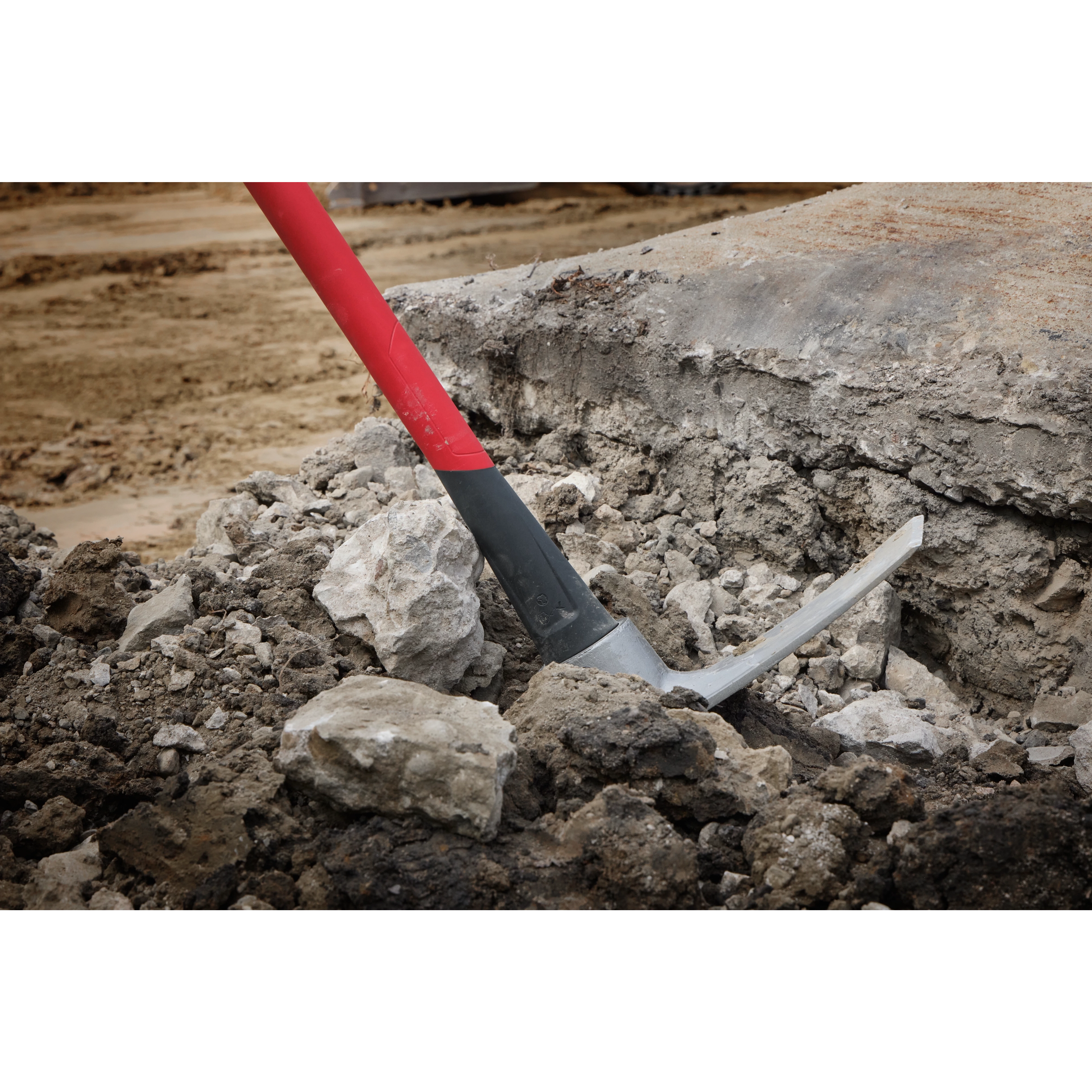 A 5lb Pick Mattock with a 36-inch handle is shown in use, breaking through hard soil and rocks. The tool's red and black handle is visible, along with the pick head firmly lodged into the ground, demonstrating its capability for heavy-duty digging and excavation tasks.
