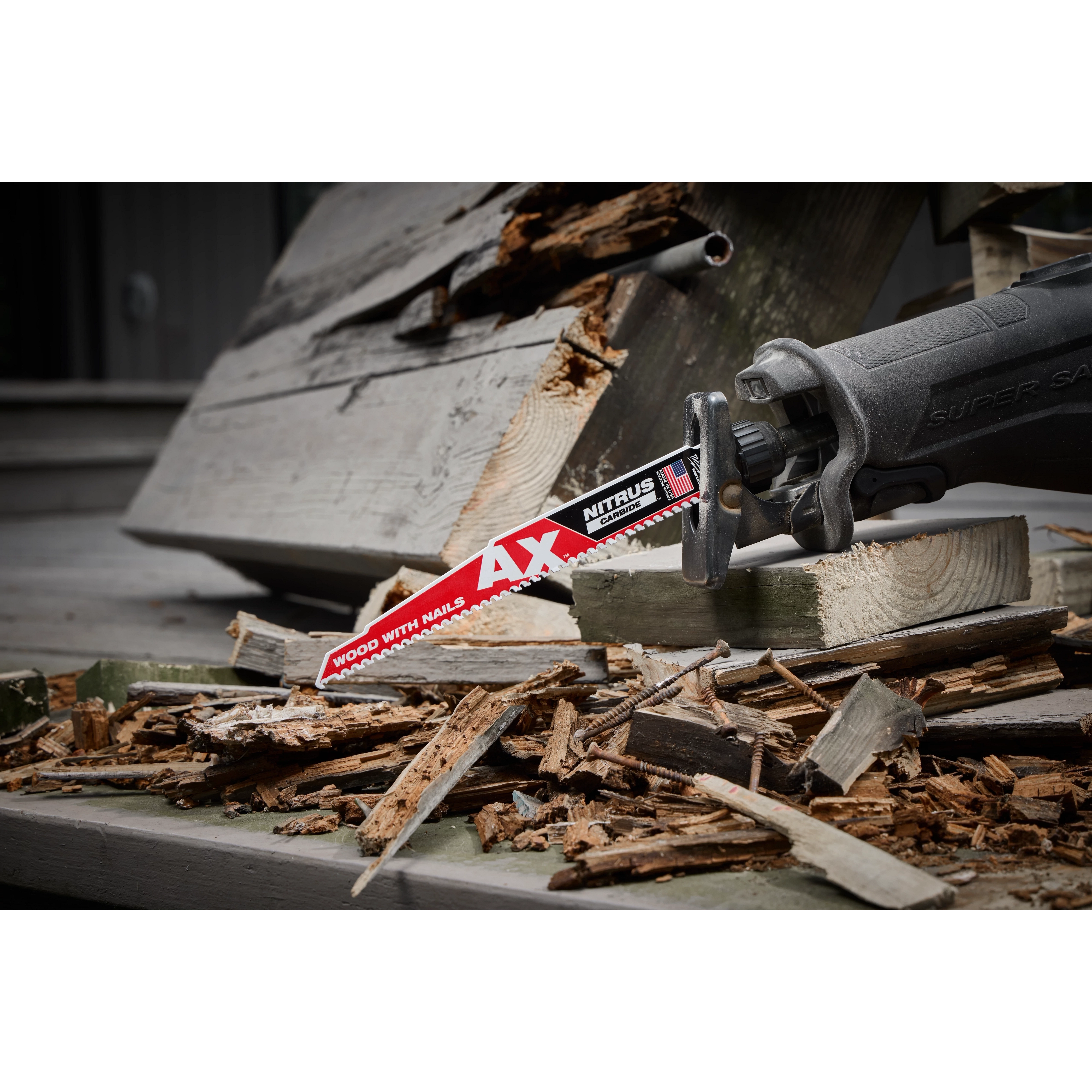 A reciprocating saw equipped with a Milwaukee Ax Nitrous Carbide Tipped Blade is shown cutting through wood and nails. The scene depicts heavy-duty demolition work, with the blade easily slicing through the debris. The saw's handle is visible, featuring an ergonomic design for user comfort and control.