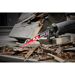 A reciprocating saw equipped with a Milwaukee Ax Nitrous Carbide Tipped Blade is shown cutting through wood and nails. The scene depicts heavy-duty demolition work, with the blade easily slicing through the debris. The saw's handle is visible, featuring an ergonomic design for user comfort and control.