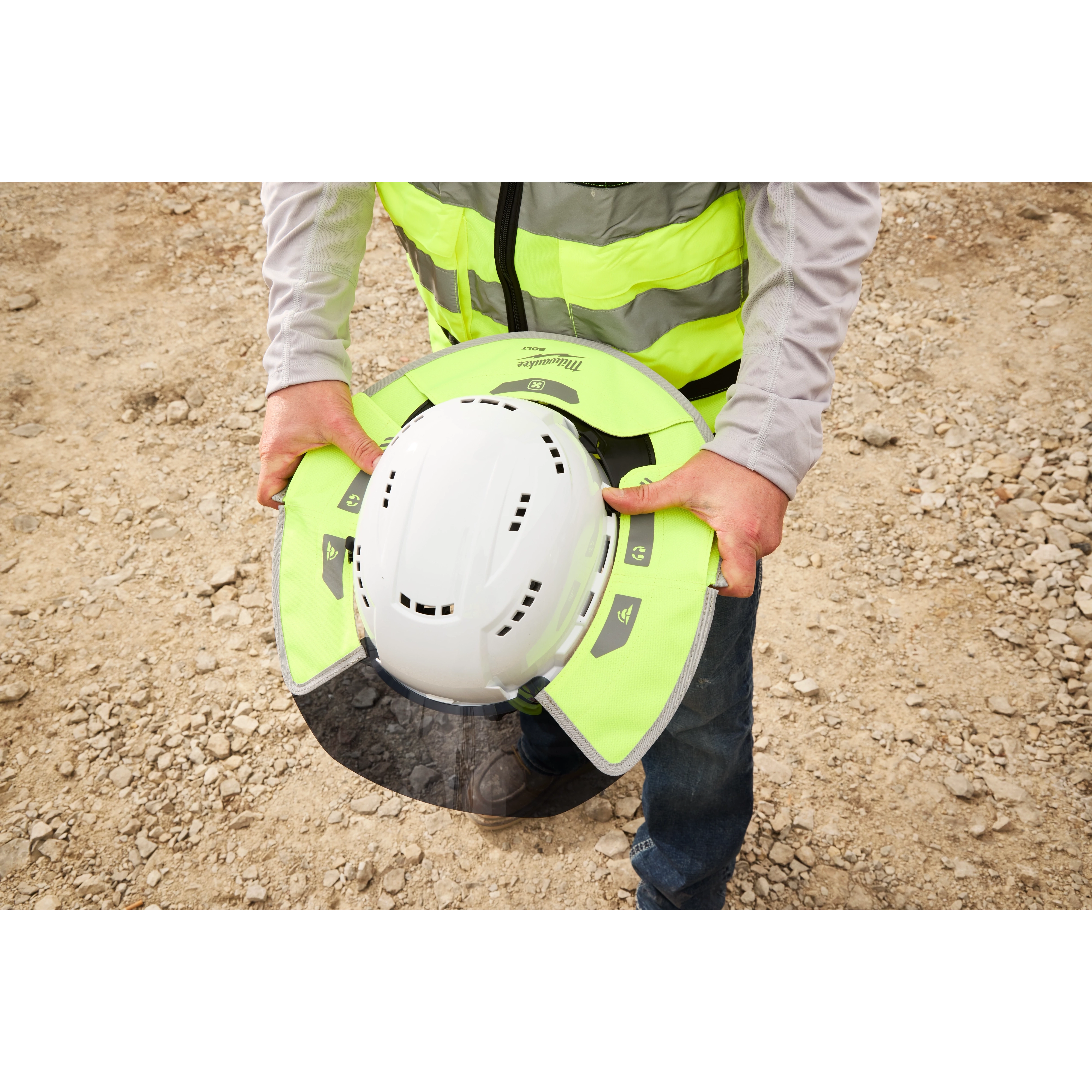 A construction worker is holding a white safety helmet equipped with a neon yellow sun shield attachment. The worker is wearing a reflective safety vest. The background shows a gravelly construction site.