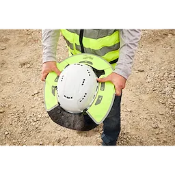 A construction worker is holding a white safety helmet equipped with a neon yellow sun shield attachment. The worker is wearing a reflective safety vest. The background shows a gravelly construction site.