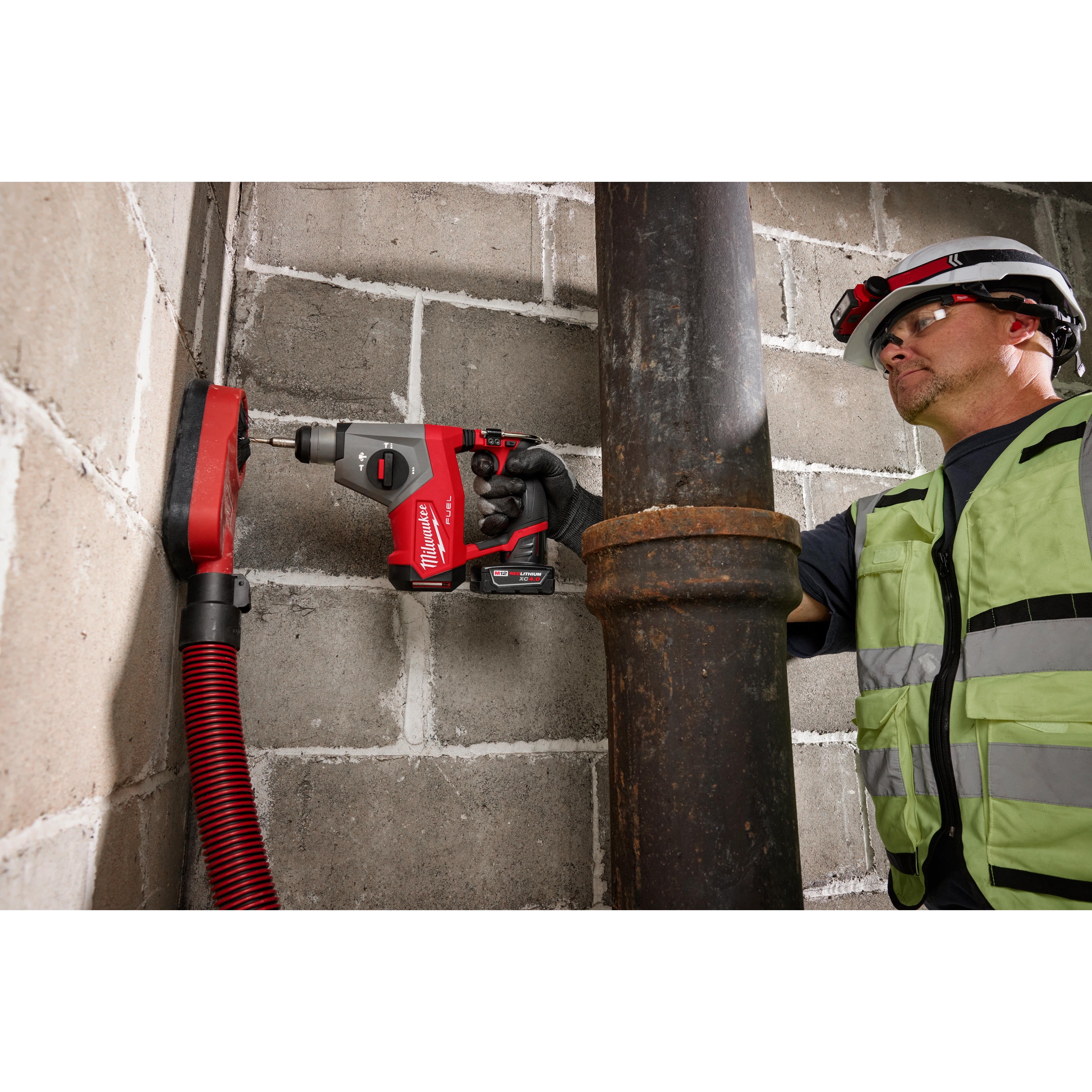 A person using an M12 FUEL™ 5/8” SDS Plus Rotary Hammer to drill into a concrete wall next to a large metal pipe. The person is wearing safety gear, including a hard hat and high-visibility vest. A red hose is connected to the drilling area for debris extraction.