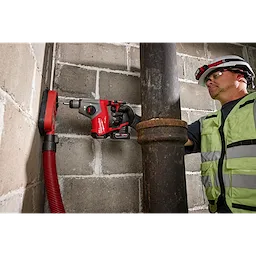 A person using an M12 FUEL™ 5/8” SDS Plus Rotary Hammer to drill into a concrete wall next to a large metal pipe. The person is wearing safety gear, including a hard hat and high-visibility vest. A red hose is connected to the drilling area for debris extraction.