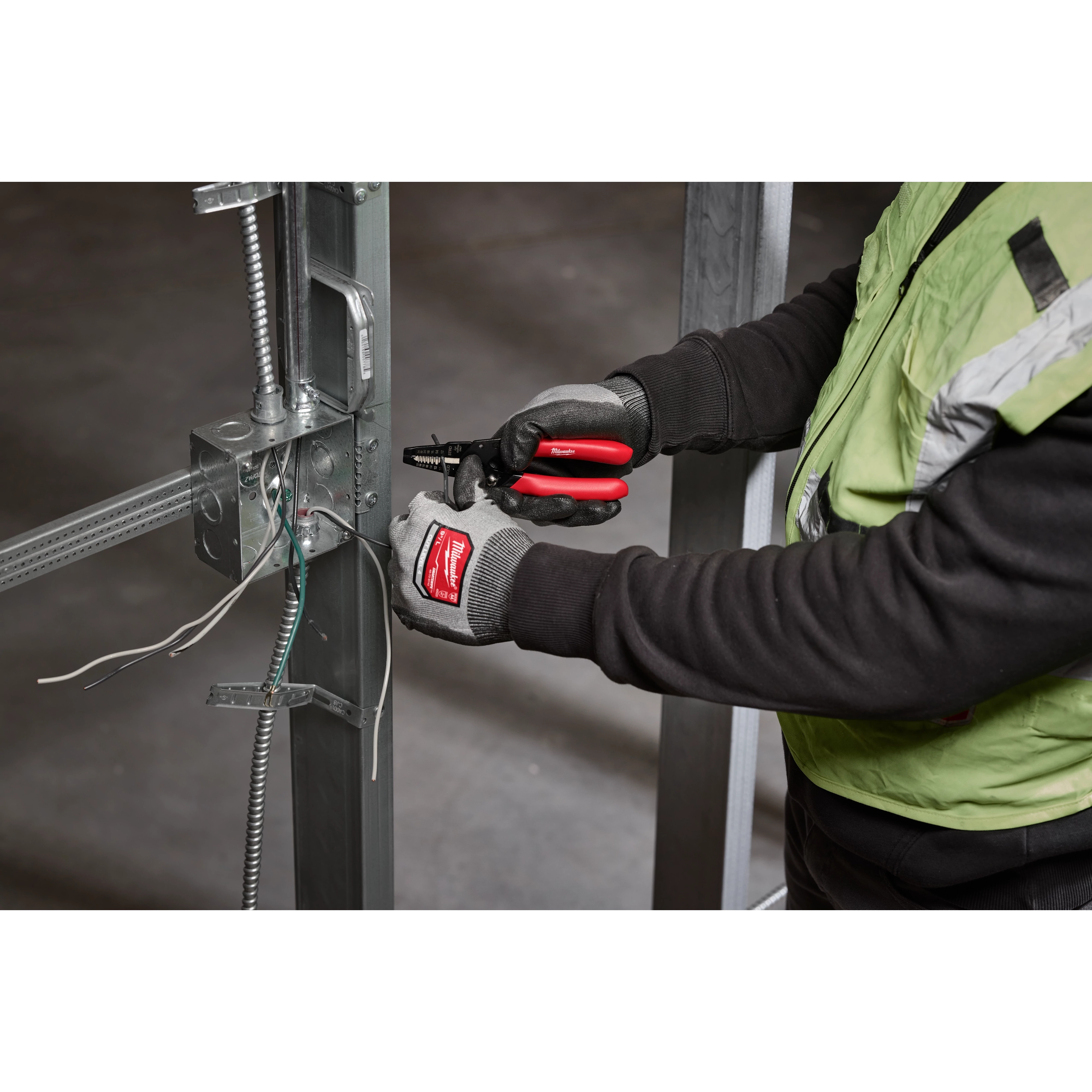 A person using the 10-24 AWG Compact Dipped Grip Wire Stripper & Cutter to strip and cut wires attached to a metal frame. The person is wearing gloves and a safety vest.