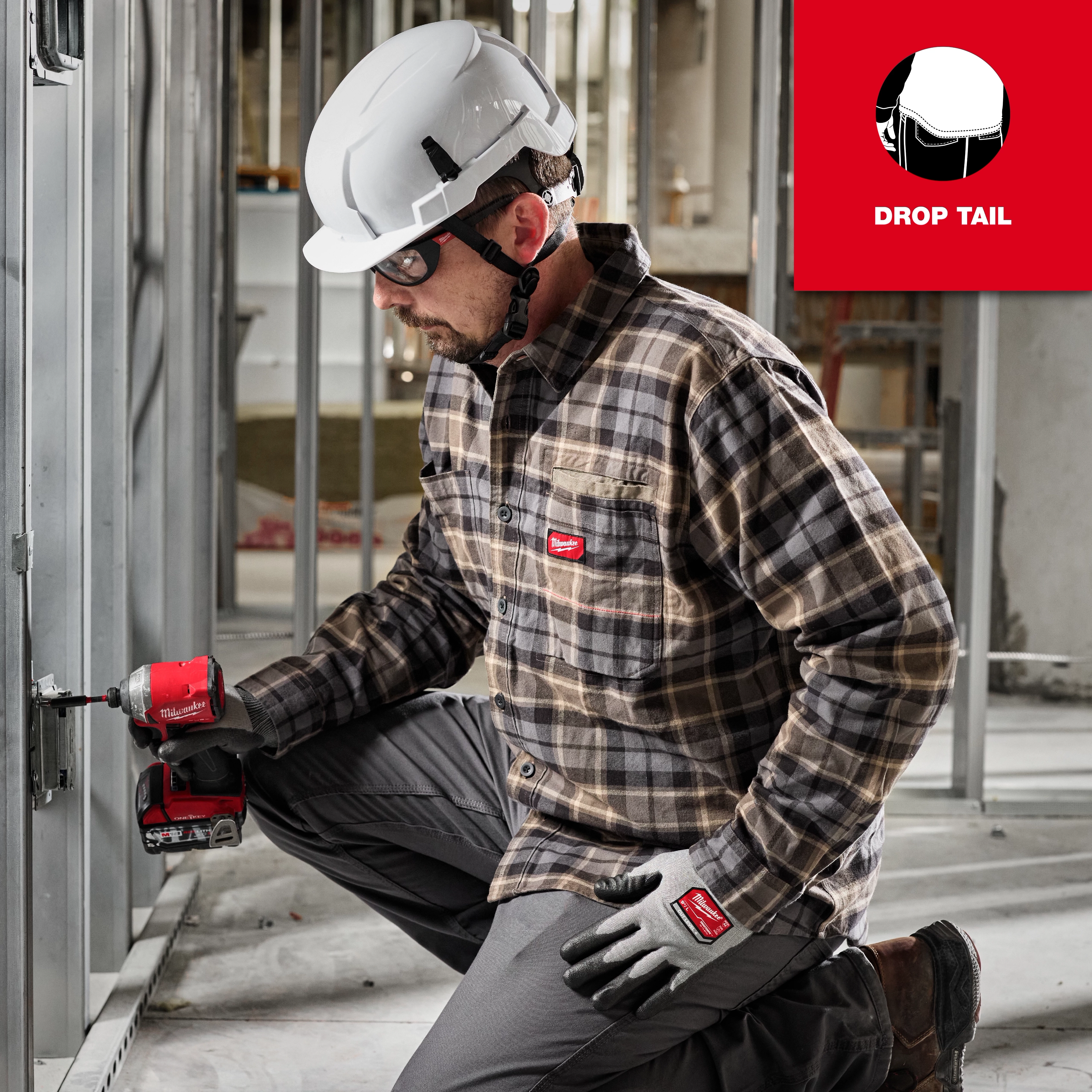 A person in a white hard hat is using a power tool while wearing a GRIDIRON Flannel Shirt, pants, and gloves with a knee pad.