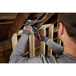 A person wearing gloves uses a 10-28 AWG Multi-Purpose Dipped Grip Wire Stripper & Cutter w/ Reinforced Head on an electrical box in an attic. The tool has red and black handles and is being used to strip wires.