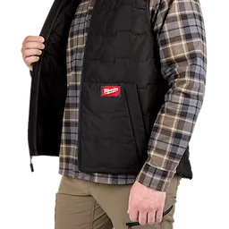 A person wears a black FREEFLEX™ Lightweight Insulated Vest over a gray and beige plaid shirt. The vest has a zipper, front pockets, and a red Milwaukee logo on the left side. The person is shown from the chest down, holding the vest open with one hand.