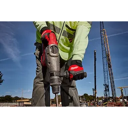 A construction worker wearing Anti-Vibration Work Gloves operates a power tool on a construction site. The worker is dressed in a reflective jacket and grey pants, and the background shows construction cranes and a clear sky.