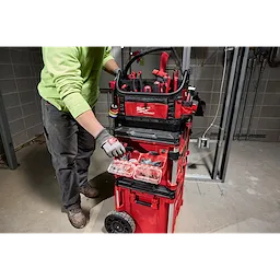 Person using PACKOUT Tool Box 2-Bin Attachment in a construction setting. The tool box is red with multiple compartments and tools.
