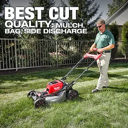 A person operates an M18 FUEL™ 21" Self-Propelled Dual Battery Mower on a lawn. The mower is red and black with a silver base. Text reads "BEST CUT QUALITY: MULCH, BAG, SIDE DISCHARGE." Trees and a fence are in the background.