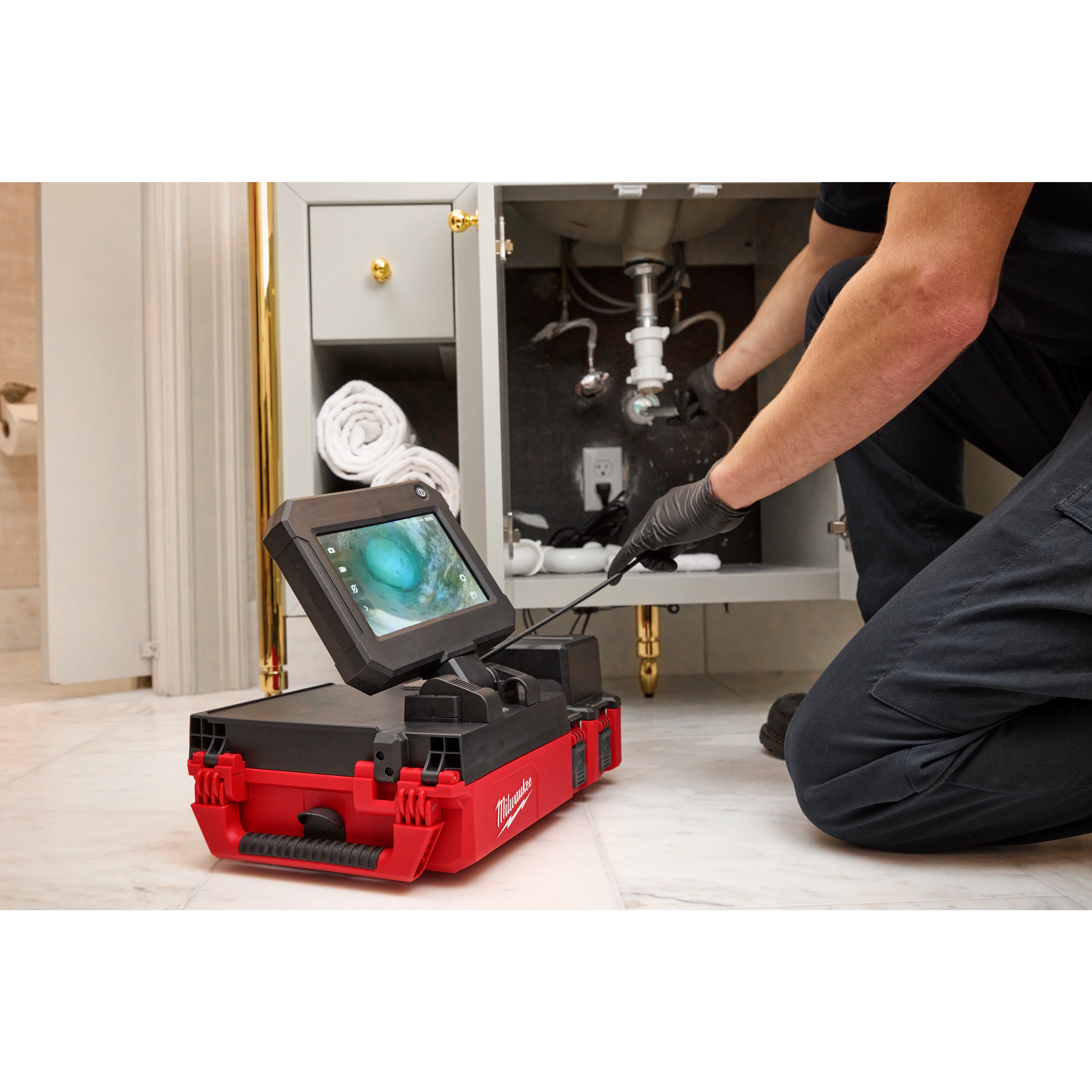A person uses the M12™ 75' Drain Camera with PACKOUT™ Compatibility to inspect pipes under a sink. The camera's monitor is placed on a red and black PACKOUT™ case, displaying a video feed. The person is kneeling on the floor, wearing gloves.
