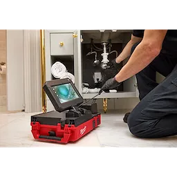 A person uses the M12™ 75' Drain Camera with PACKOUT™ Compatibility to inspect pipes under a sink. The camera's monitor is placed on a red and black PACKOUT™ case, displaying a video feed. The person is kneeling on the floor, wearing gloves.