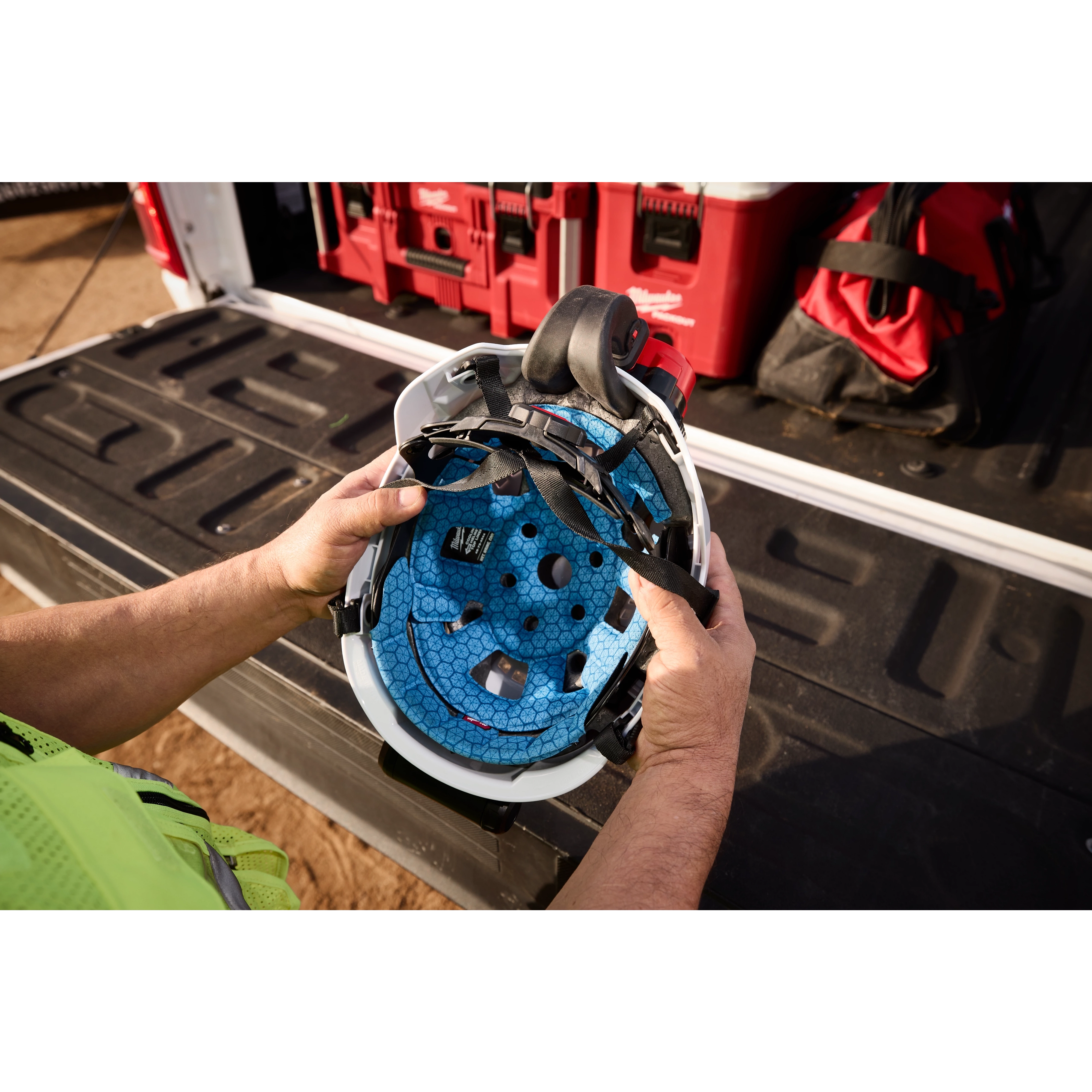 Hands holding a white hard hat with the BOLT REDLITHIUM USB Cooling Fan attached inside, against a background of red toolboxes and road case.