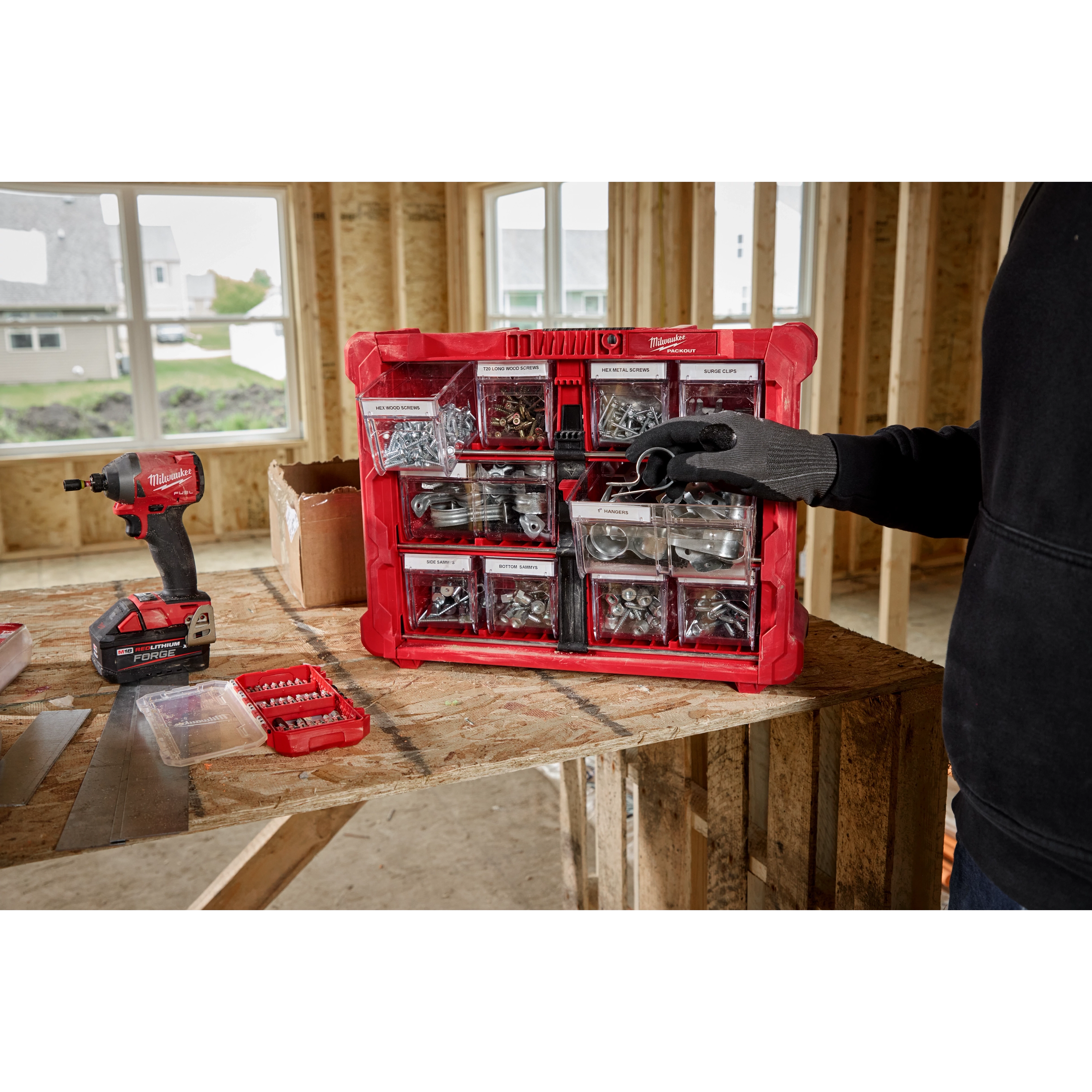 A person in a black hoodie and work glove is organizing a red Milwaukee toolkit containing various screws and metal fasteners. The toolkit is placed on a wooden table in a construction site, with an electric drill and other tools visible nearby.