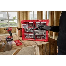 A person in a black hoodie and work glove is organizing a red Milwaukee toolkit containing various screws and metal fasteners. The toolkit is placed on a wooden table in a construction site, with an electric drill and other tools visible nearby.
