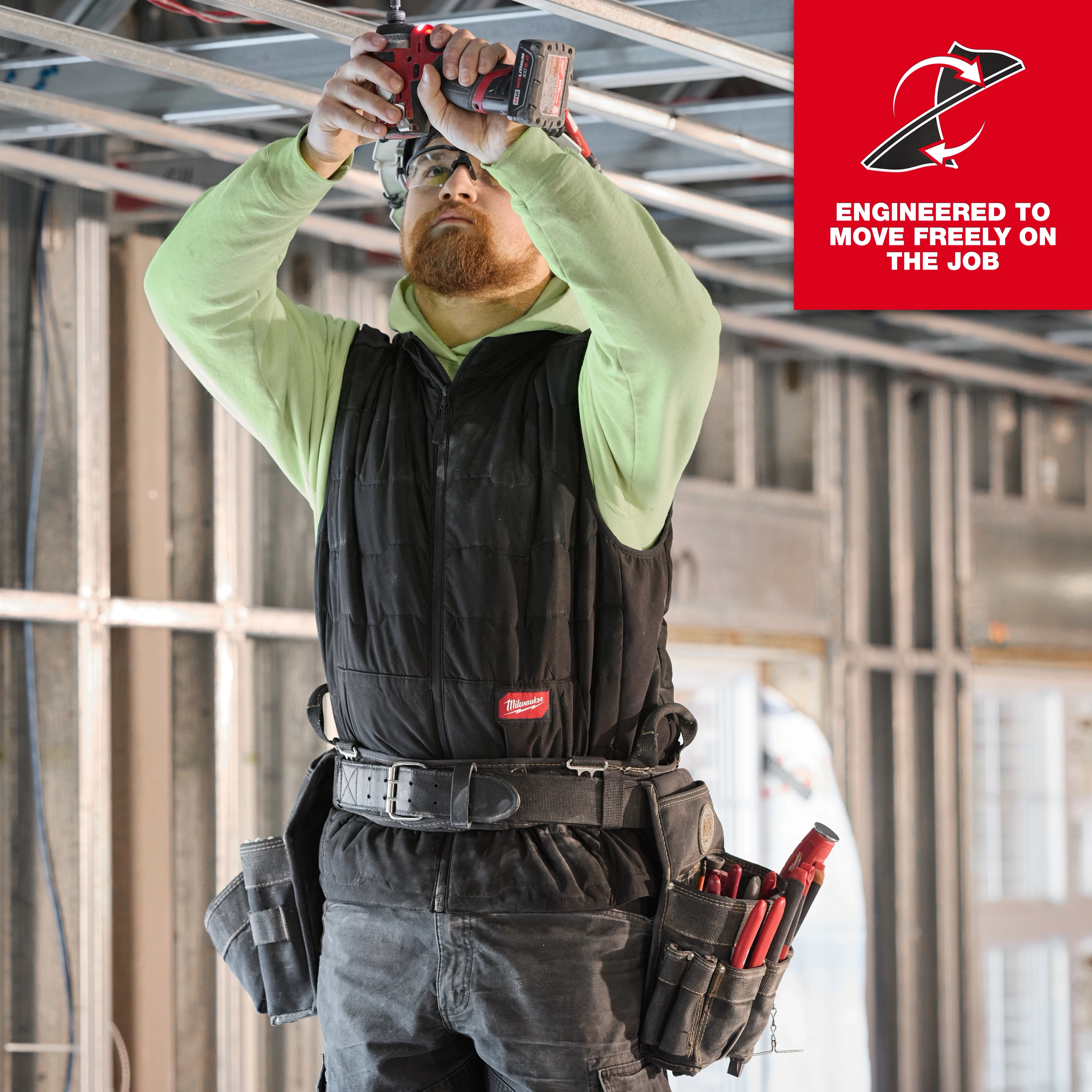 A person wearing the FREEFLEX™ Lightweight Insulated Vest is working on a construction site. The vest is black and designed for mobility, with multiple tool belts and pouches attached. The background includes metal frame structures under construction. A slogan reads "ENGINEERED TO MOVE FREELY ON THE JOB."
