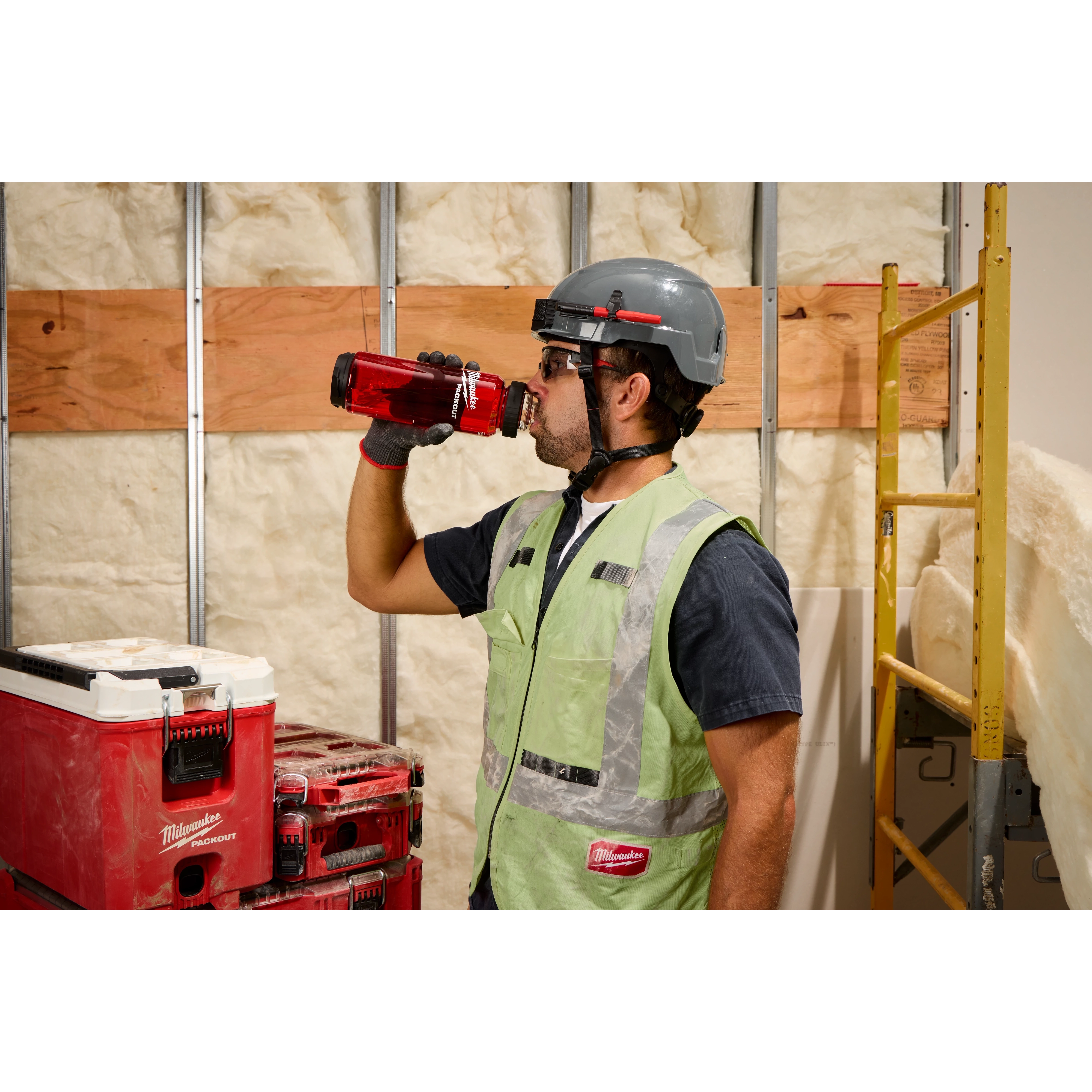 A person wearing a safety vest, gloves, and a hard hat drinks from a red PACKOUT™ 34oz Bottle with Chug Lid. They stand next to red and white PACKOUT™ storage units in a construction site with insulation and building materials in the background.