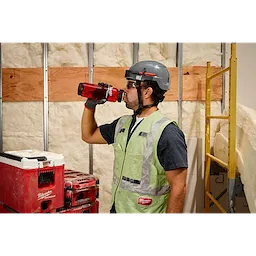 A person wearing a safety vest, gloves, and a hard hat drinks from a red PACKOUT™ 34oz Bottle with Chug Lid. They stand next to red and white PACKOUT™ storage units in a construction site with insulation and building materials in the background.