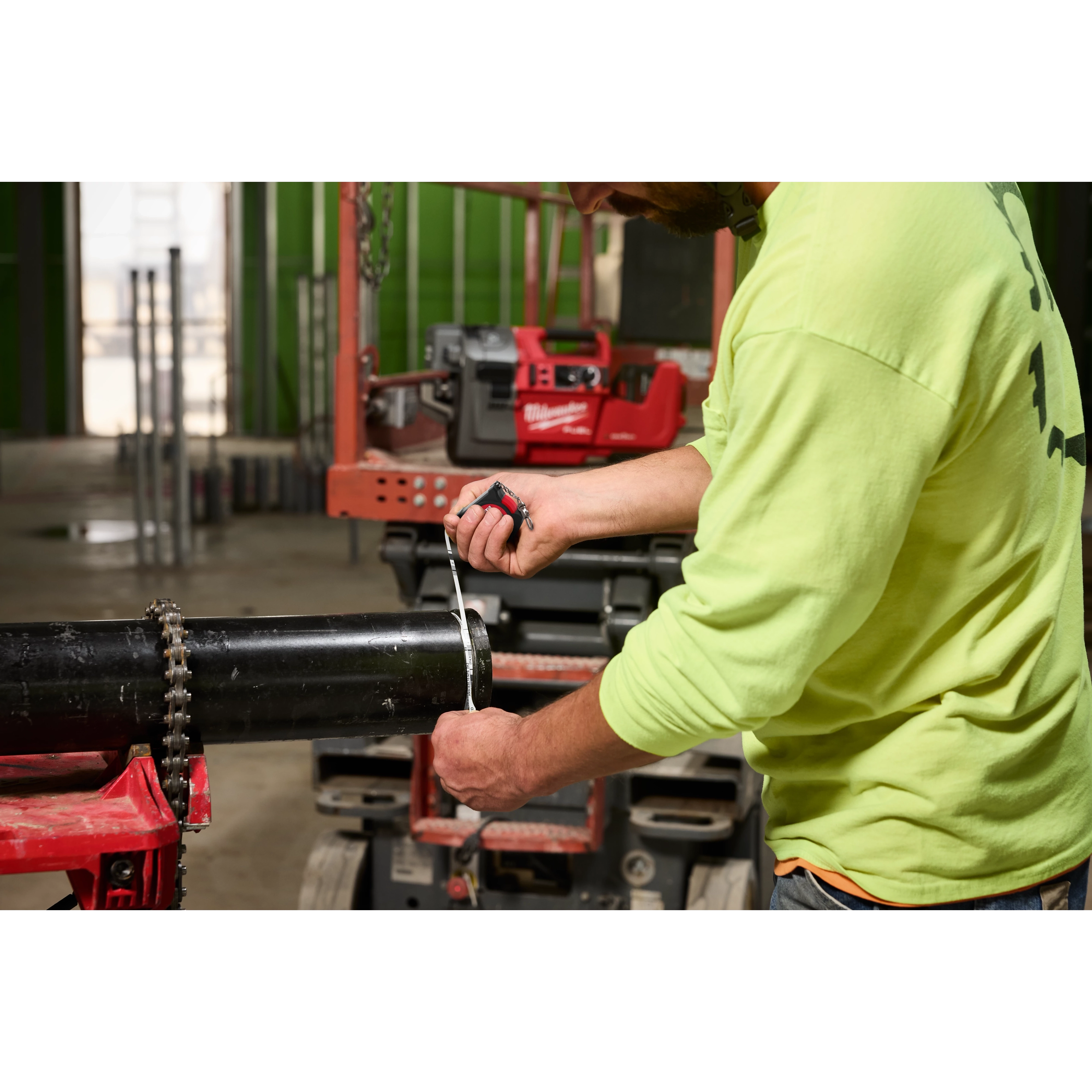 A person in a yellow shirt measures a black pipe using a measuring tape. The M18 FUEL™ RINGER™ Roll Groover for 2” – 6” Sch 10/40 tool is visible in the background, mounted on a workbench in a construction setting.