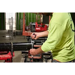 A person in a yellow shirt measures a black pipe using a measuring tape. The M18 FUEL™ RINGER™ Roll Groover for 2” – 6” Sch 10/40 tool is visible in the background, mounted on a workbench in a construction setting.