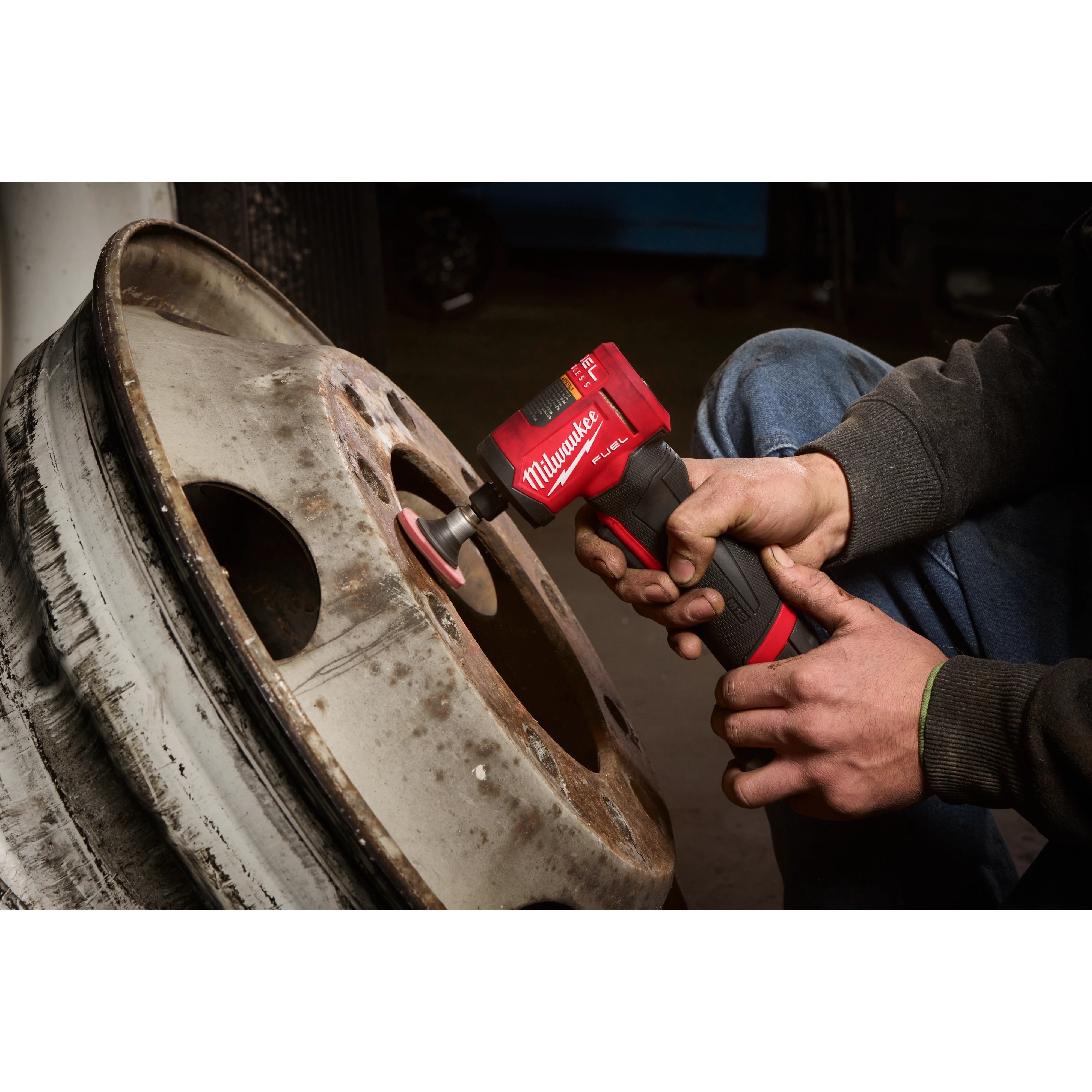 A person uses the M12 FUEL™ 1/4" Right Angle Die Grinder to clean a rusty wheel rim. The red and black tool is being held with both hands, with the attachment in contact with the metal surface.