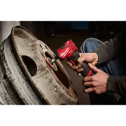 A person uses the M12 FUEL™ 1/4" Right Angle Die Grinder to clean a rusty wheel rim. The red and black tool is being held with both hands, with the attachment in contact with the metal surface.