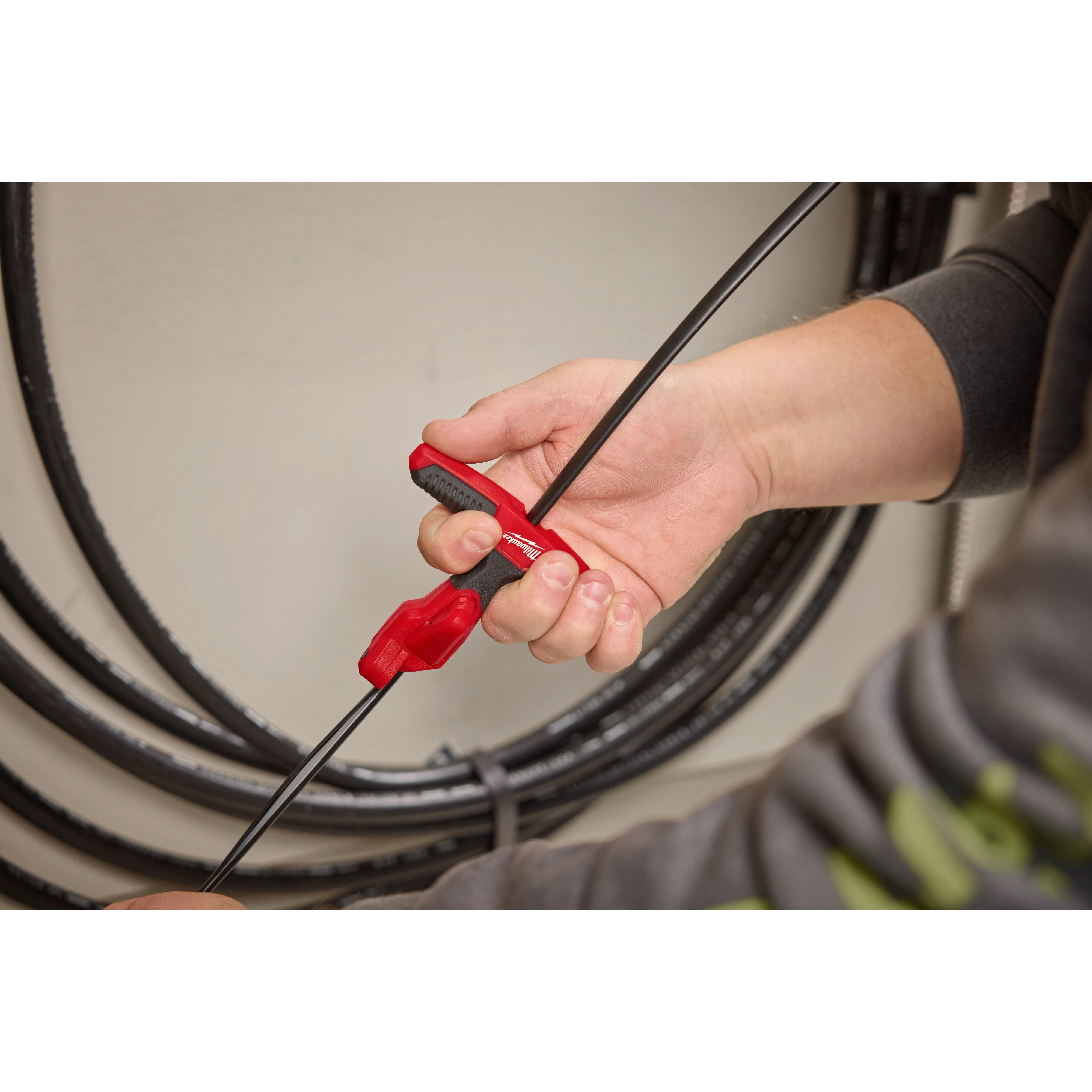 A person is holding a Fiber Optic Drop Cable Slitter, a tool with a red handle, to cut through a black fiber optic cable, with coiled cables visible in the background.