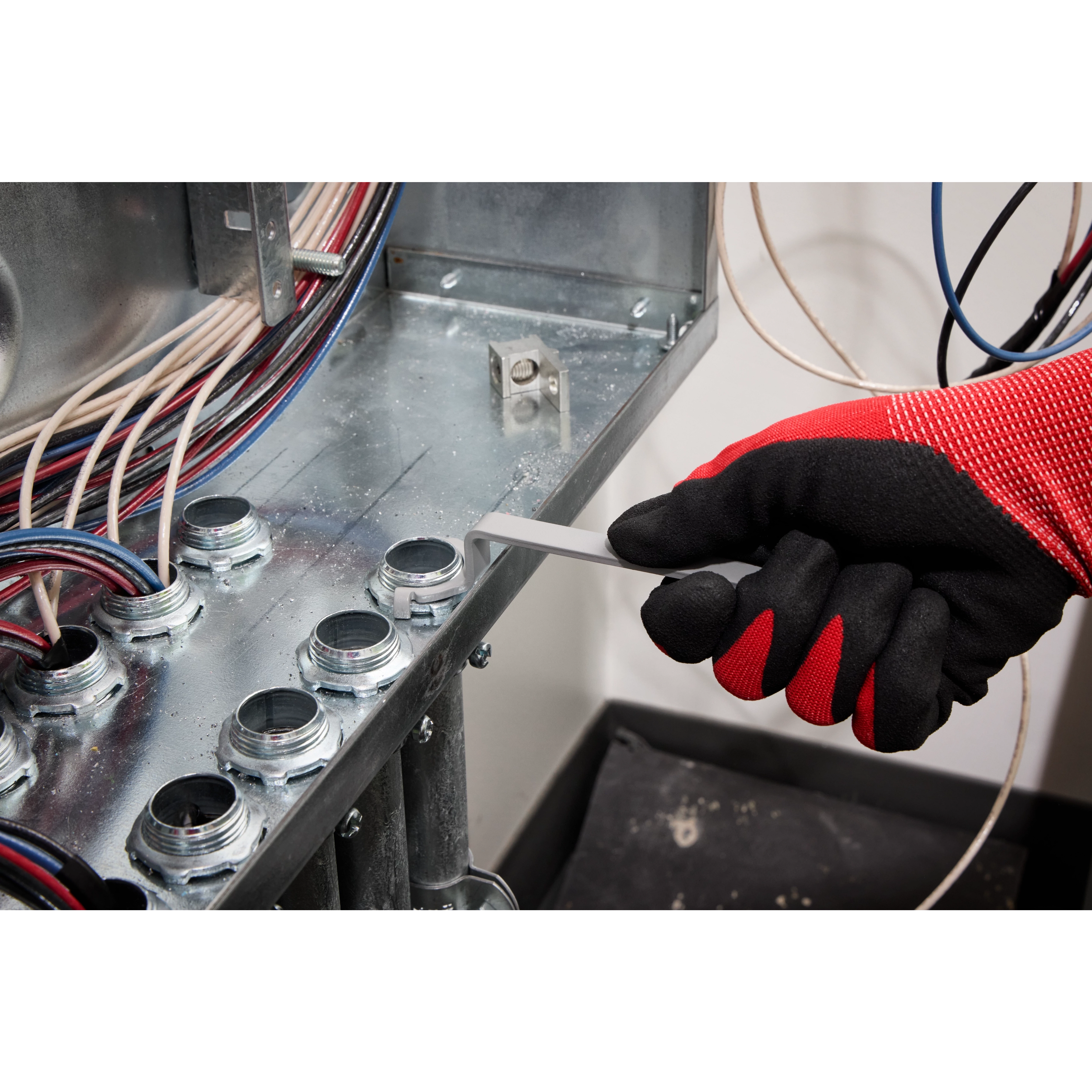 A gloved hand uses a wrench from the 3 PC Locknut Wrench Set to tighten a locknut inside an electrical panel. Multiple wires and locknuts are visible inside the panel, and the hand is wearing black and red gloves for protection. The wrenches are designed for electrical work.