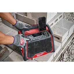 Worker wearing gloves adjusts the M18™ BLUETOOTH® Jobsite Radio + Charger, which is designed with a rugged exterior and large speaker. An upright smartphone is docked on top of the radio. The radio is placed on stacked concrete blocks.