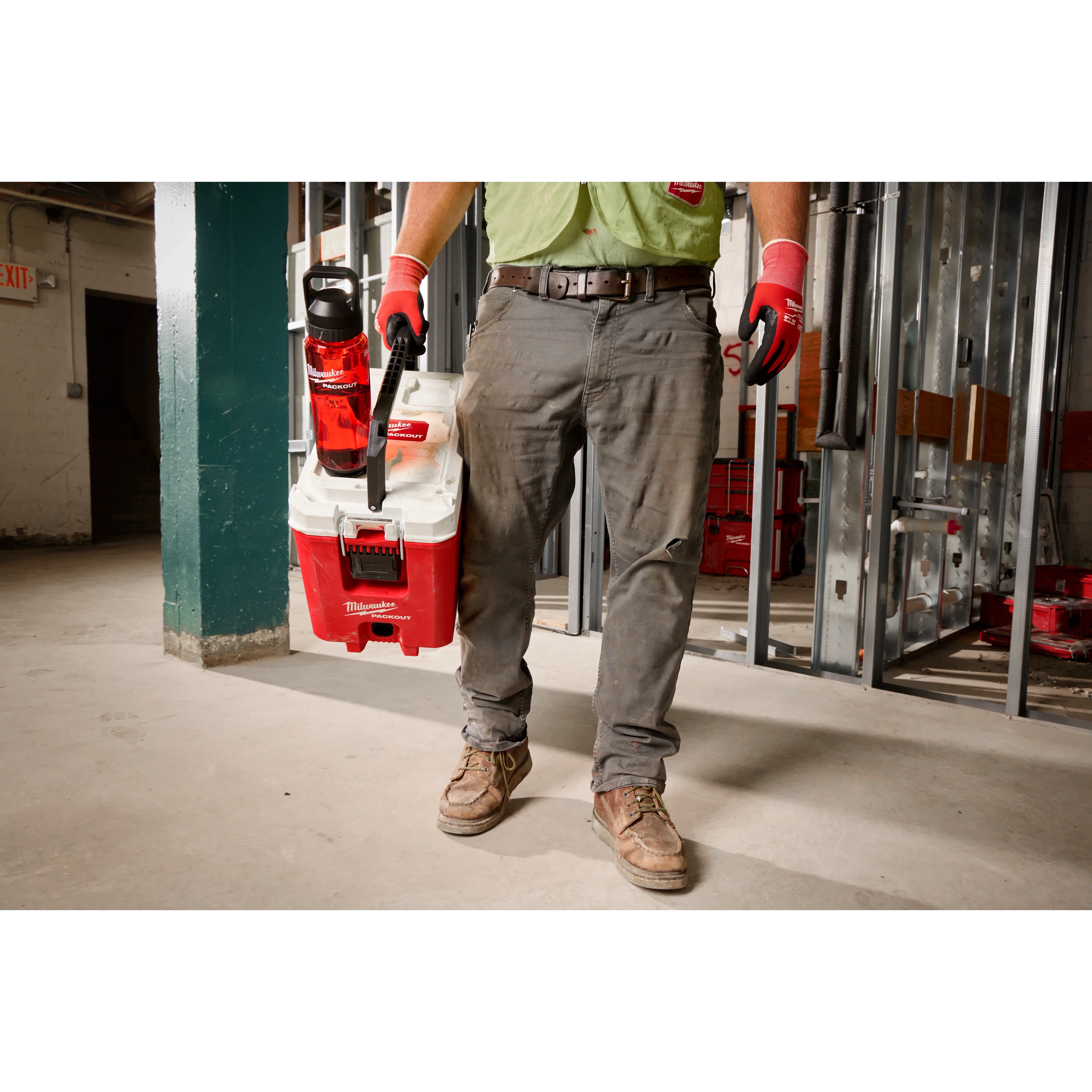 A person wearing work gloves and boots is holding a white and red Milwaukee tool bag. Attached to the bag is a PACKOUT™ 34oz Bottle with Chug Lid, which is red with a black lid. The setting appears to be an industrial or construction site, with metal framing and tools in the background.