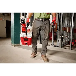 A person wearing work gloves and boots is holding a white and red Milwaukee tool bag. Attached to the bag is a PACKOUT™ 34oz Bottle with Chug Lid, which is red with a black lid. The setting appears to be an industrial or construction site, with metal framing and tools in the background.