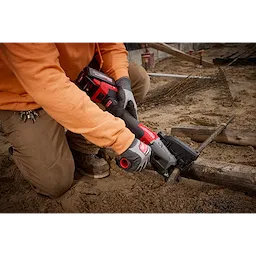 A person is using an M18 FUEL™ #10 (1-1/4") Rebar Cutter to cut a piece of rebar on a construction site. The tool is black and red, and the person is wearing an orange hoodie, brown pants, and work gloves.