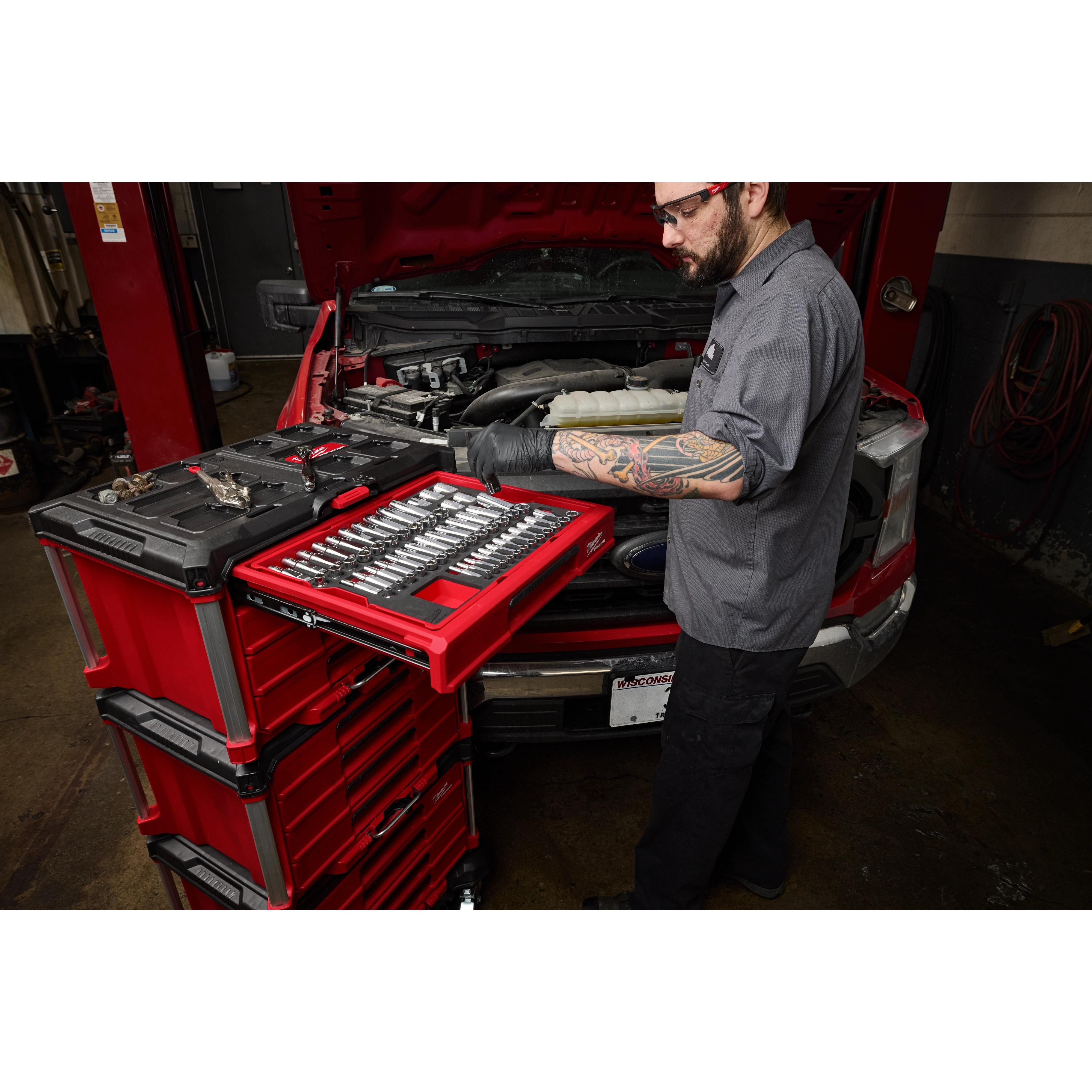 A mechanic works on a car engine using a 366pc. Master Mechanics Hand Tool Set with PACKOUT™ Drawers and Dolly. The tool set is organized in red drawers, with various tools visible on the workbench.