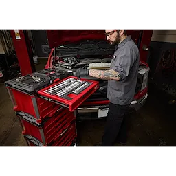 A mechanic works on a car engine using a 366pc. Master Mechanics Hand Tool Set with PACKOUT™ Drawers and Dolly. The tool set is organized in red drawers, with various tools visible on the workbench.