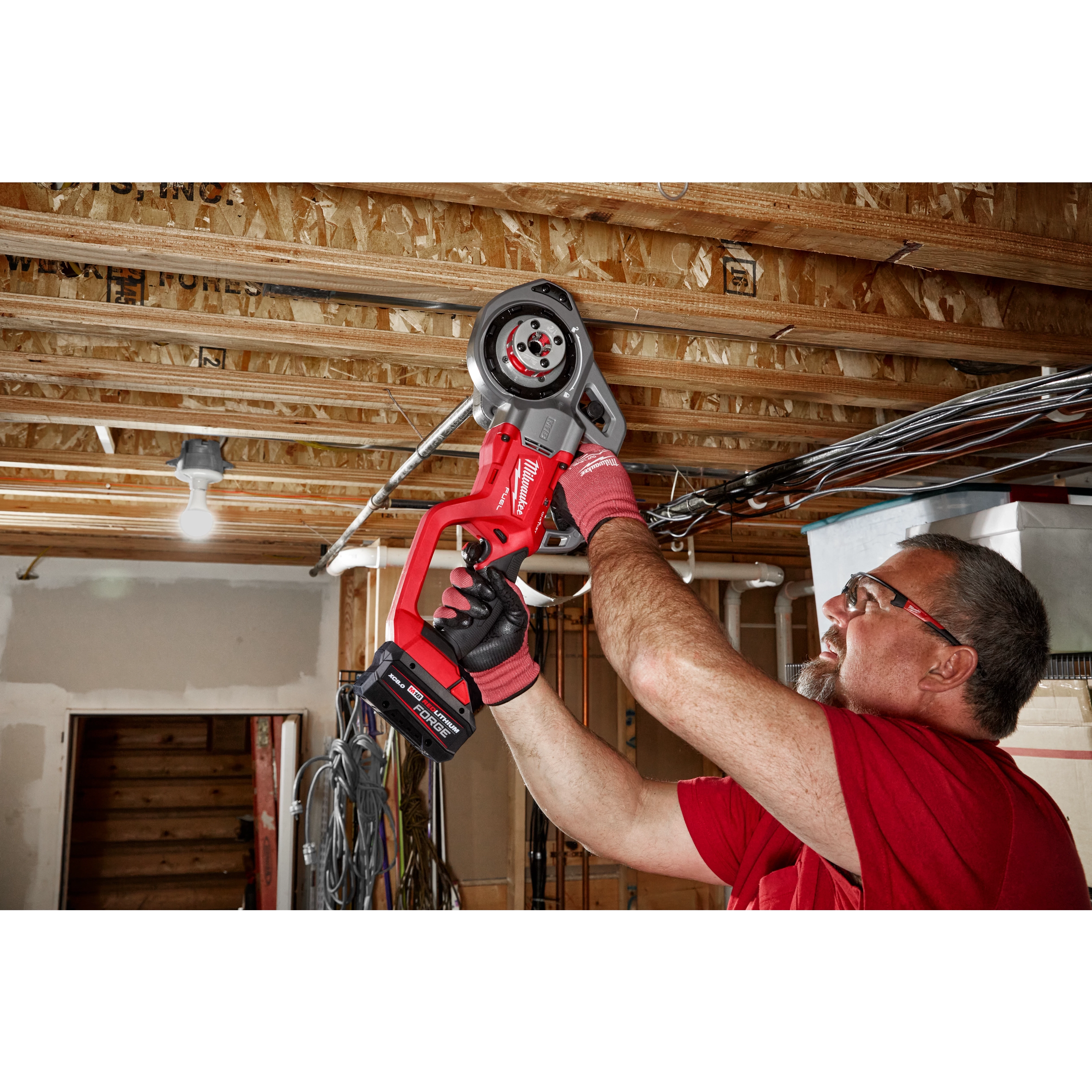 A person uses the M18 FUEL™ Compact Threader with One-Key™ to work on pipes installed in a ceiling. The tool is red and black, and the setting is an unfinished basement or construction site.