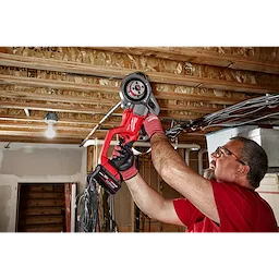 A person uses the M18 FUEL™ Compact Threader with One-Key™ to work on pipes installed in a ceiling. The tool is red and black, and the setting is an unfinished basement or construction site.