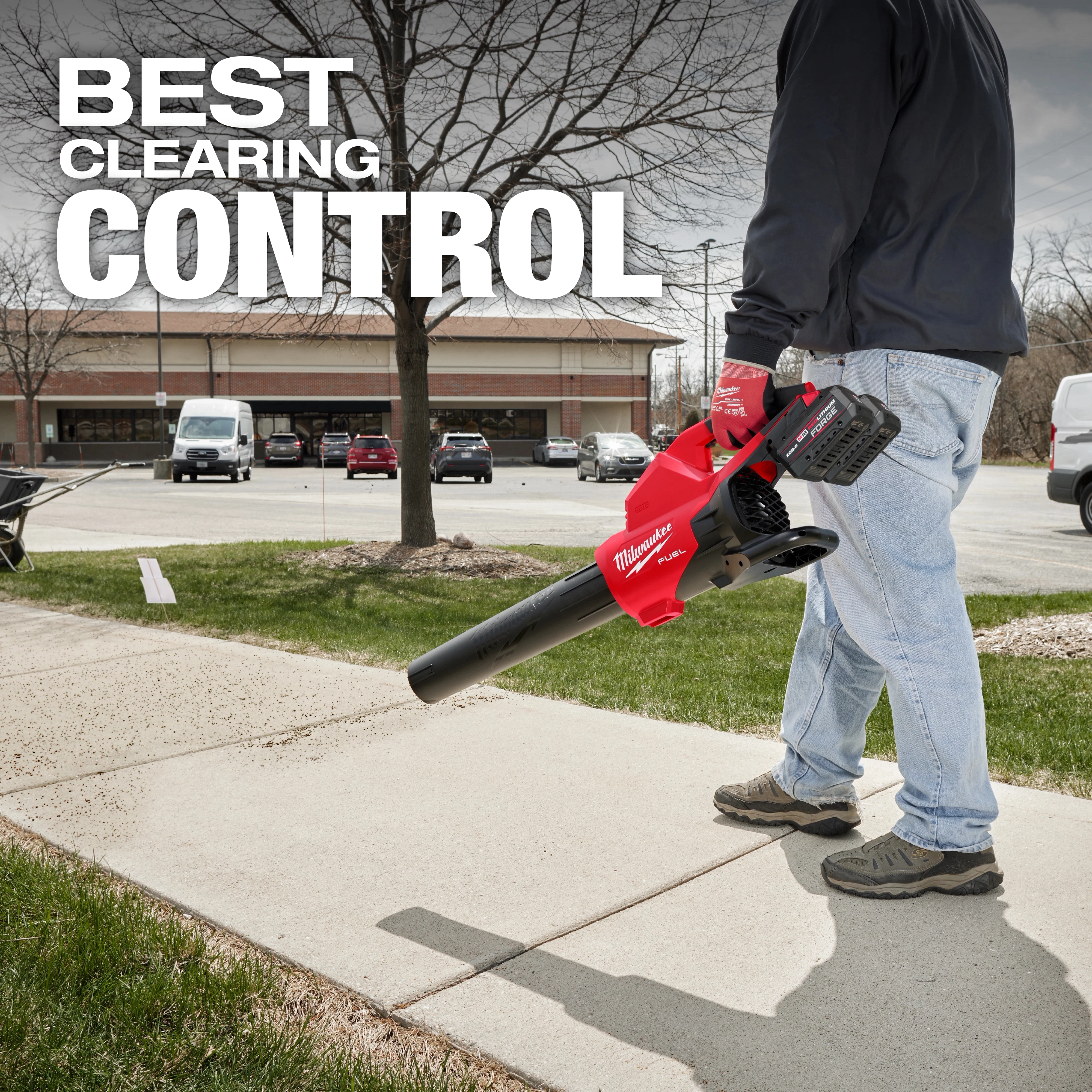 A person uses the M18 FUEL™ Dual Battery Blower to clear a sidewalk. The blower is red and black. The background shows a parking lot with cars, trees, and a building. The text "BEST CLEARING CONTROL" is displayed at the top.